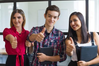 Portrait of successful young students showing thumbs up. Group of two Caucasian girls and boy looking at camera and smiling at college. Success and education concept